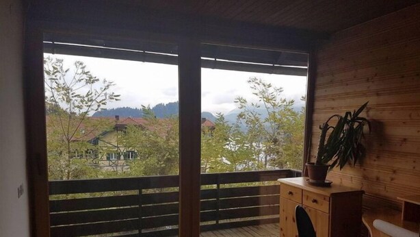 A room with a wooden floor, a large sliding glass door leading to a balcony, a wooden cabinet with a plant, and a chair. - Beautiful 3-bedroom apartment with lake view (Radovljica)