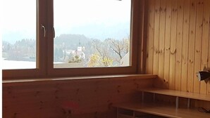 A bedroom with a bed, a bedside table, a small stool, a wooden wardrobe, and a window with a view of a church and hills. - Beautiful 3-bedroom apartment with lake view (Radovljica)