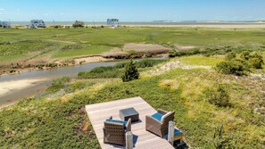 Une terrasse en bois, meublée de meubles en osier, qui offre une vue sur un paysage côtier avec une rivière et des maisons situées au loin.