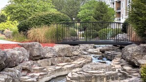 A landscaped area with a stone waterfall, a metal bridge, and a red mulch border. - Branson | Pool & Mountains | Scenic Ozark Stay (Banson)