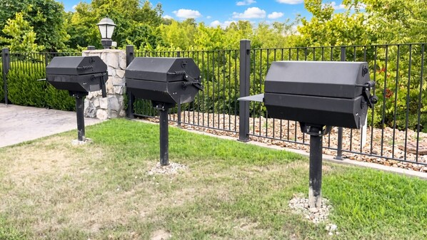 Trois barbecues d’extérieur sont disposés sur une pelouse, avec un mur de pierre et une clôture métallique en arrière-plan.