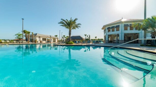 Piscina azul cristalina con borde curvo, rodeada de palmeras y un edificio con múltiples ventanas.