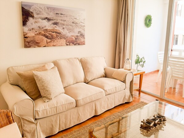 Un divano beige con cuscini decorati, un tavolino da caffè in vetro e un quadro raffigurante le onde sulla parete.