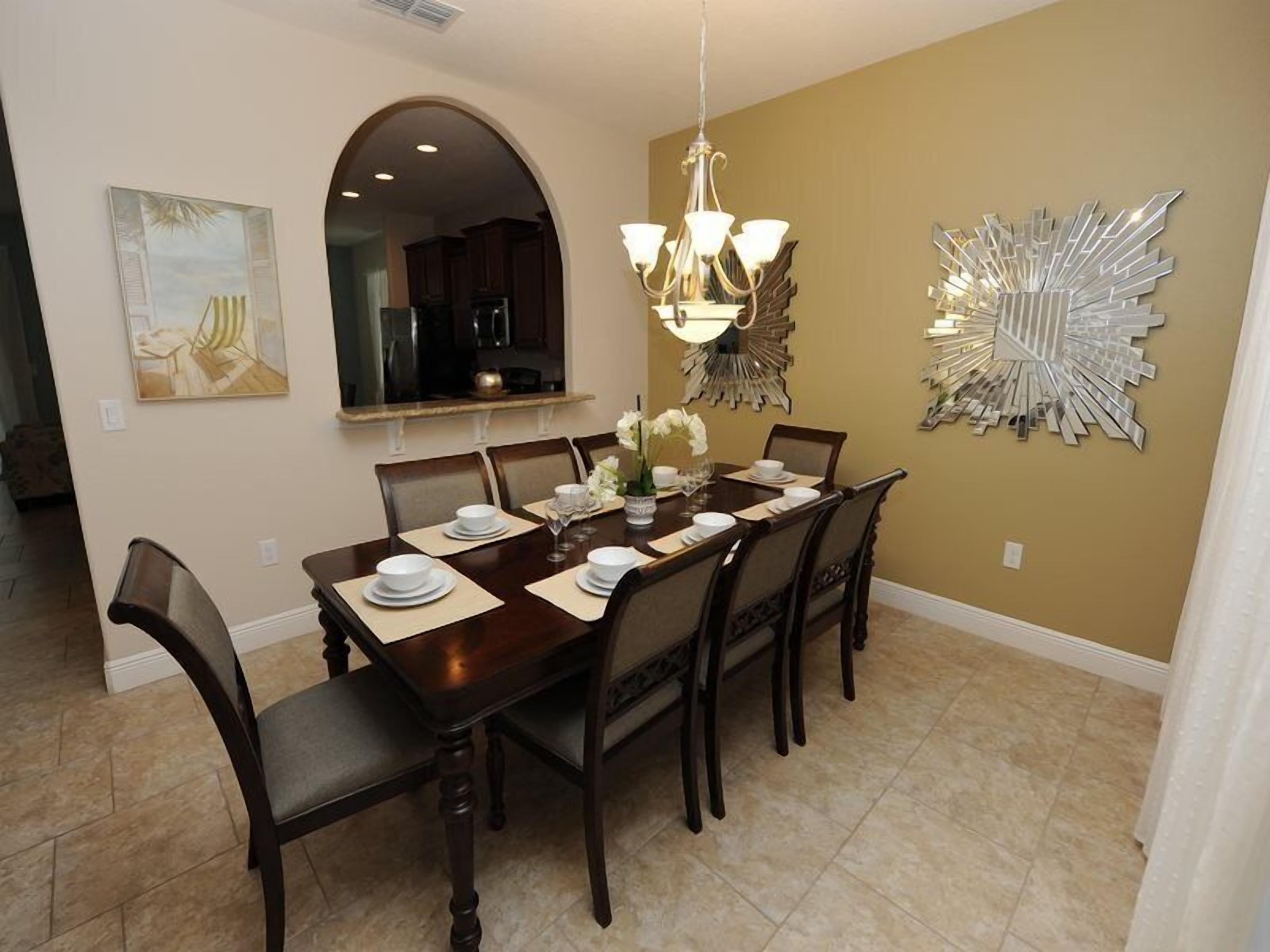 A dining room with a long table set for six, a chandelier, and a decorative mirror on the wall.