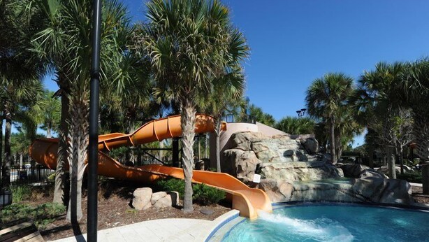 Un espace piscine doté d’un toboggan aquatique, de palmiers et d’une cascade artificielle en roches.