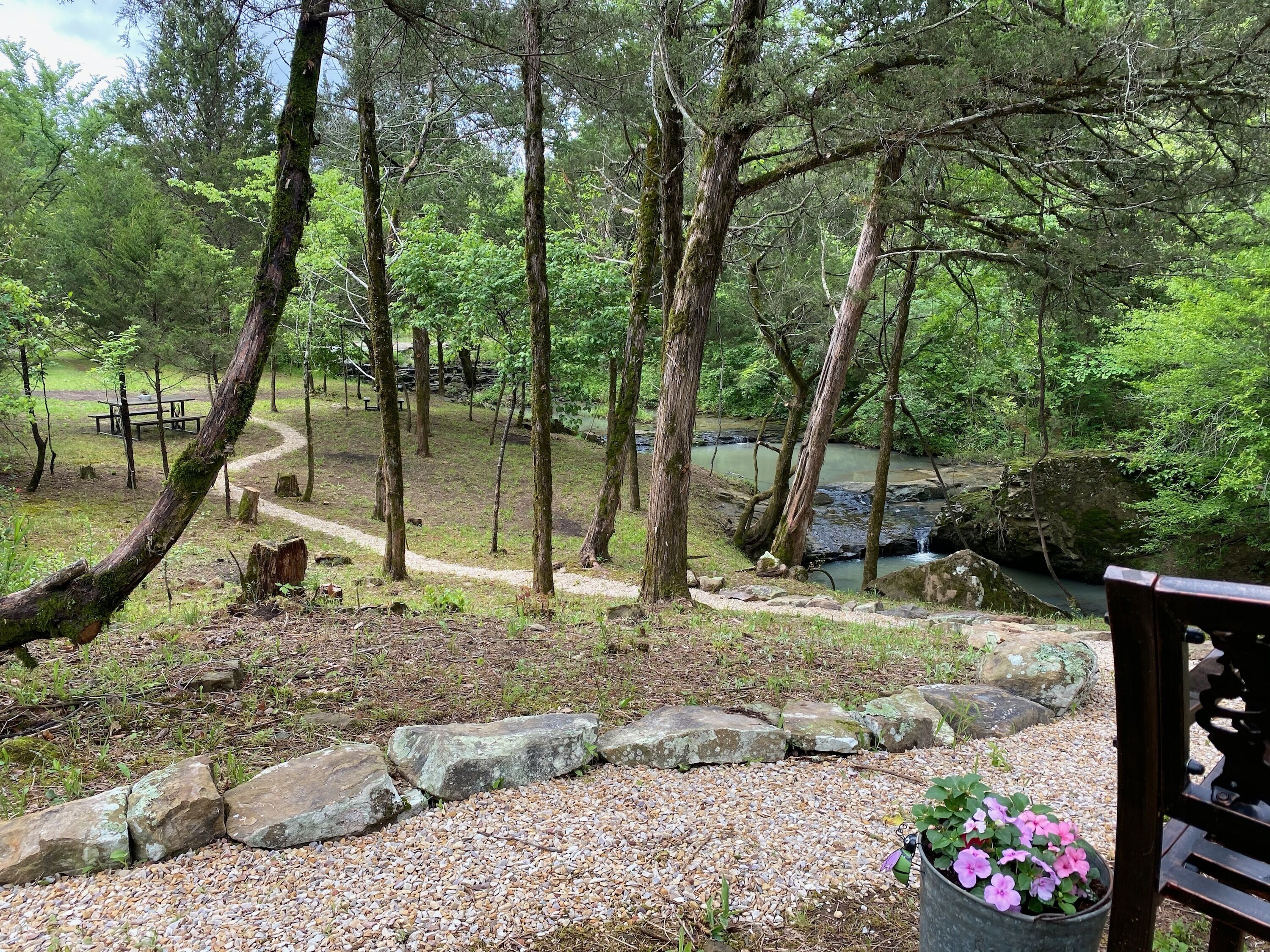 A forested area with a gravel path, a picnic table, and a small stream.