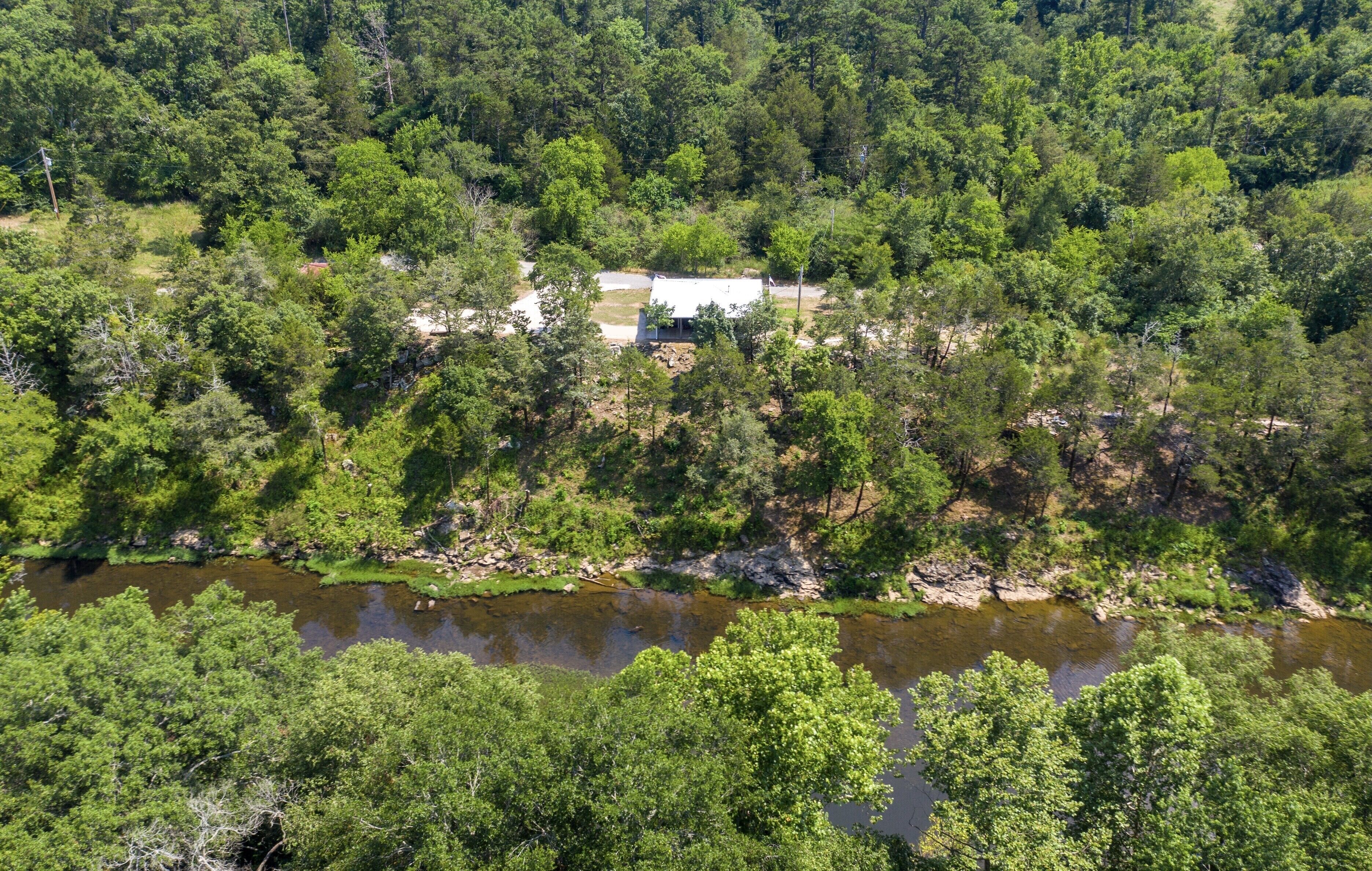 Vue aérienne d’une propriété entourée d’une forêt dense, avec un petit bâtiment et une rivière au premier plan.