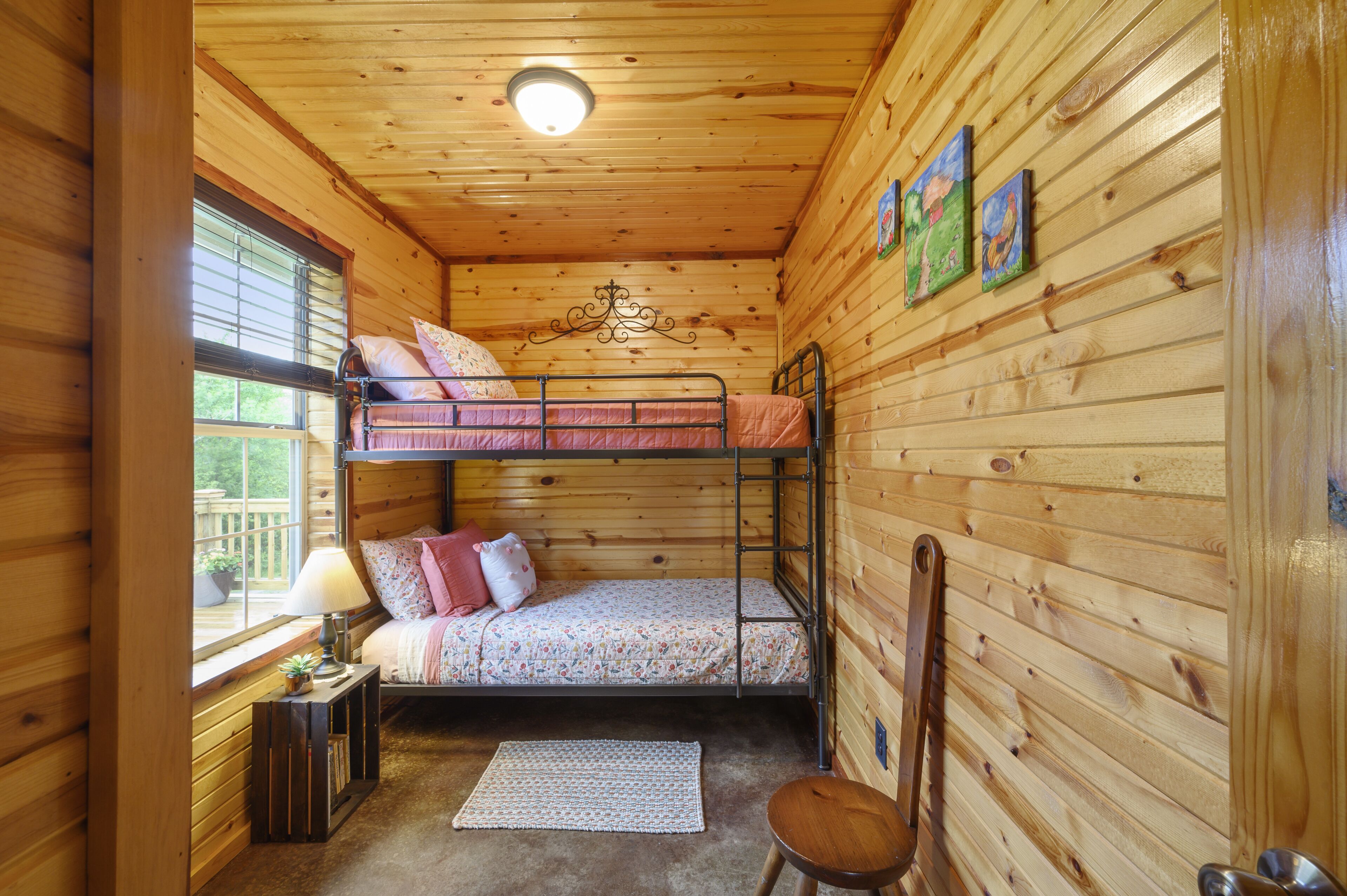 A cozy cabin room with a bunk bed, a small table with a lamp, a chair, and a window with blinds.