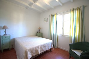 Un dormitorio con cama, mesita de noche, lámpara, silla, ventana con cortinas y un cuadro en la pared.