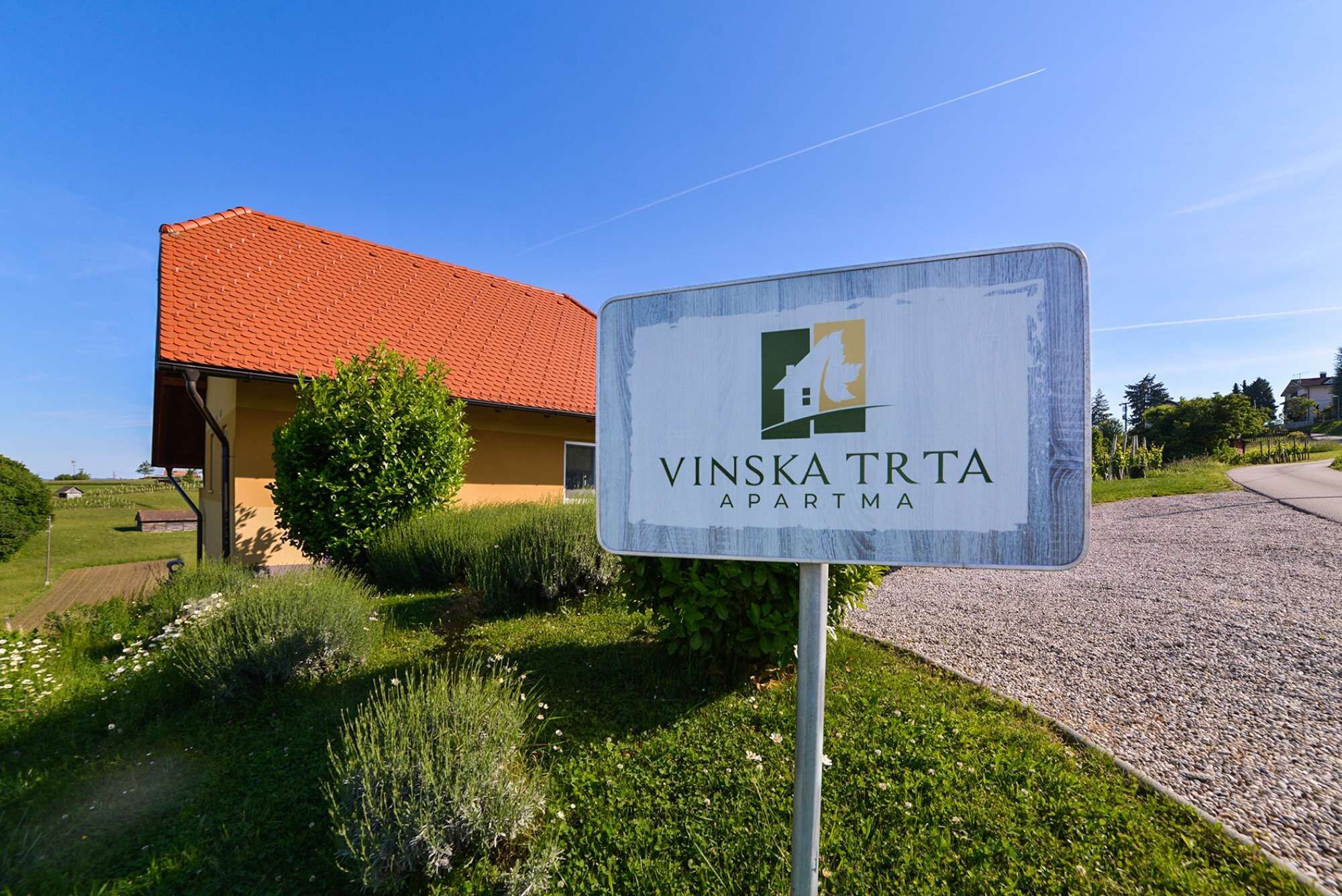 A sign for Vinska Trta apartment with a clear view of a building with a red roof and a well-maintained lawn.