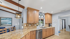 A kitchen with wooden cabinets, a stainless steel dishwasher, granite countertops, and a hanging light fixture. - Sunny Cape Coral Pool Home (Cape Coral)