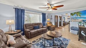 A living room with a brown leather sofa, a wooden coffee table, and a ceiling fan. There is a large window with blue curtains, a lamp on a side table, and a painting of a beach scene on the wall. - Sunny Cape Coral Pool Home (Cape Coral)