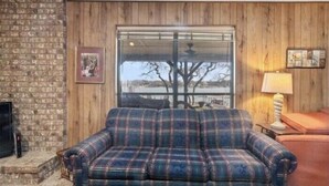 A cozy living room with a plaid sofa, a fireplace, and a window with a view of trees and a building. - The Lake Shack-----Water Front!!! (Brownwood)