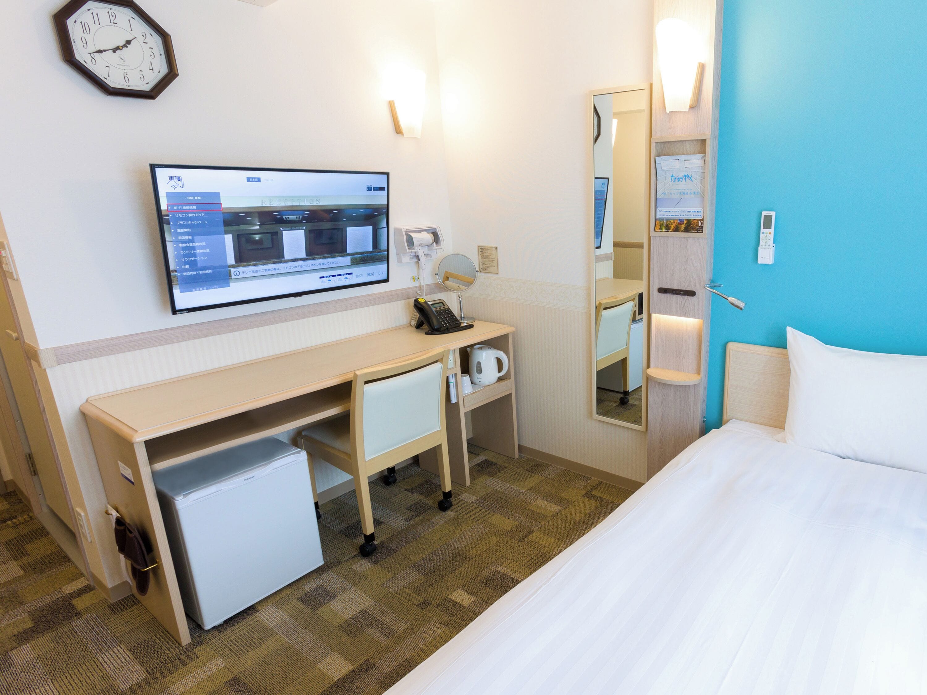 Habitación de hotel con cama, escritorio con computadora, refrigerador pequeño, espejo y un reloj en la pared.