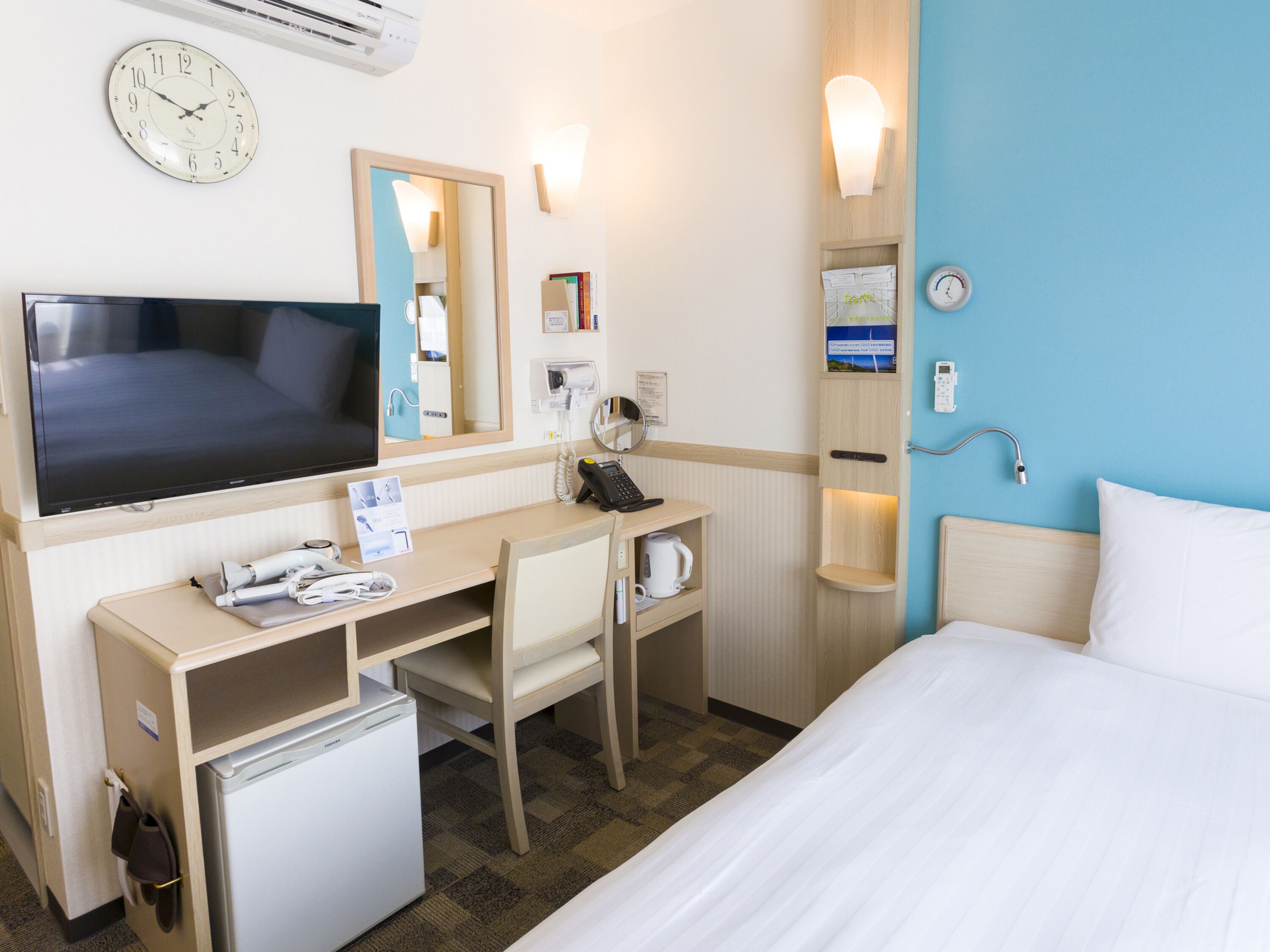 A hotel room with a bed, a desk with a computer, a television, a refrigerator, a microwave, a coffee maker, a hairdryer, a mirror, a clock, and a shower.
