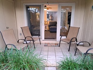 Une terrasse avec deux chaises et une petite table, une porte-fenêtre coulissante donnant sur un salon, et un paillasson orné d’un motif de plage.