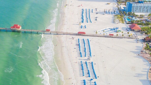 An aerial view of a beach with a long pier extending into the water, lined with beach umbrellas and a hotel with multiple buildings.