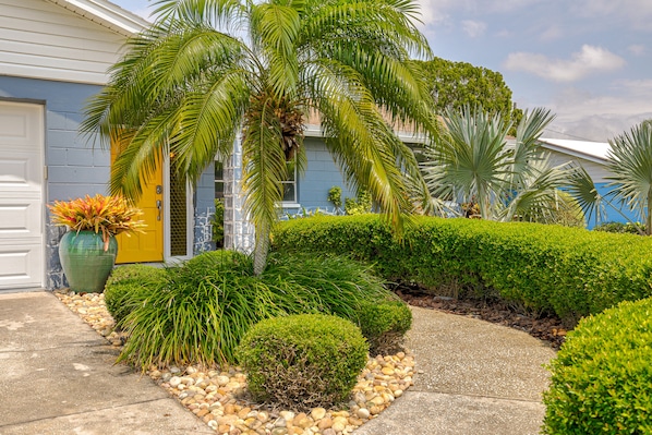 A house with a yellow door, surrounded by well-maintained landscaping including palm trees, shrubs, and a concrete pathway.