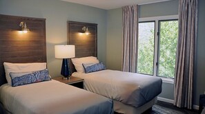 Two adjacent beds with matching headboards and bedside tables, each with a lamp, offer a comfortable retreat with a view of the surrounding greenery.