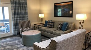 Relax in a comfortable seating area with a sofa, armchair, and ottoman, surrounded by a large painting and a window with curtains.