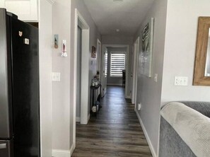 A narrow hallway with a black refrigerator on the left, a gray couch on the right, and a wooden floor.