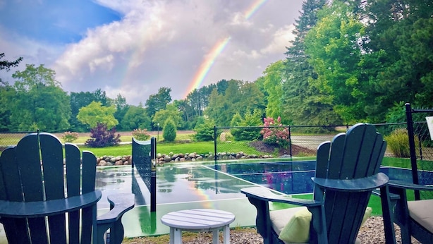 Een buitenzitgedeelte met Adirondack-stoelen en een kleine tafel, met uitzicht op een zwembad en een weelderig groen landschap met een regenboog aan de hemel.