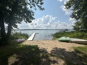 Ein sandiger Strand mit einer anlegenden Mole in einem ruhigen See, umgeben von Bäumen und mit Kajaks ausgestattet.