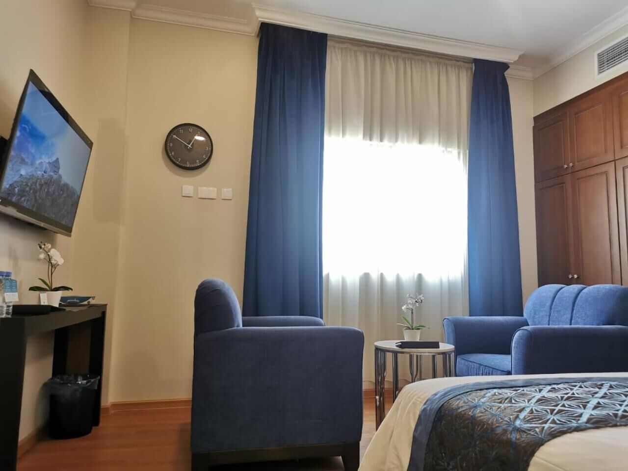 Une chambre d’hôtel avec un lit, deux fauteuils, une petite table, une horloge et un téléviseur fixé au mur.