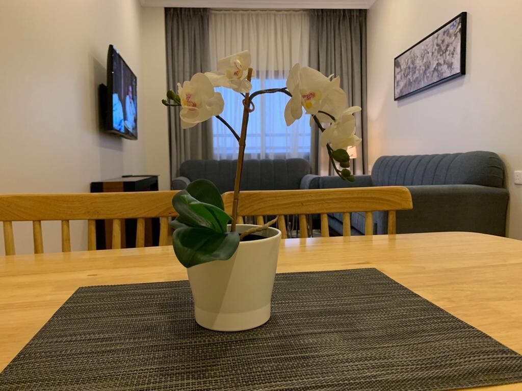 Ett vardagsrum med ett matbord i trä, en orkidé i en kruka, en grå soffa och en tv på väggen.