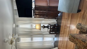 A room with a wooden floor, a patterned sofa, a small table with a lamp, a wooden cabinet, and a ceiling fan. -  Elevated Apt steps from beach. Furnished Parking Utilities, TV Internet Include (Tybee Island)
