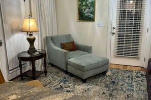 A room with a grey sofa, a small round table with a lamp, a patterned rug, and a door with blinds.