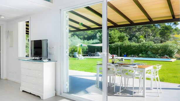 Eine moderne Küche mit Fernseher, ein Essbereich mit weißen Stühlen und eine Glasdoor zu einem Poolbereich.