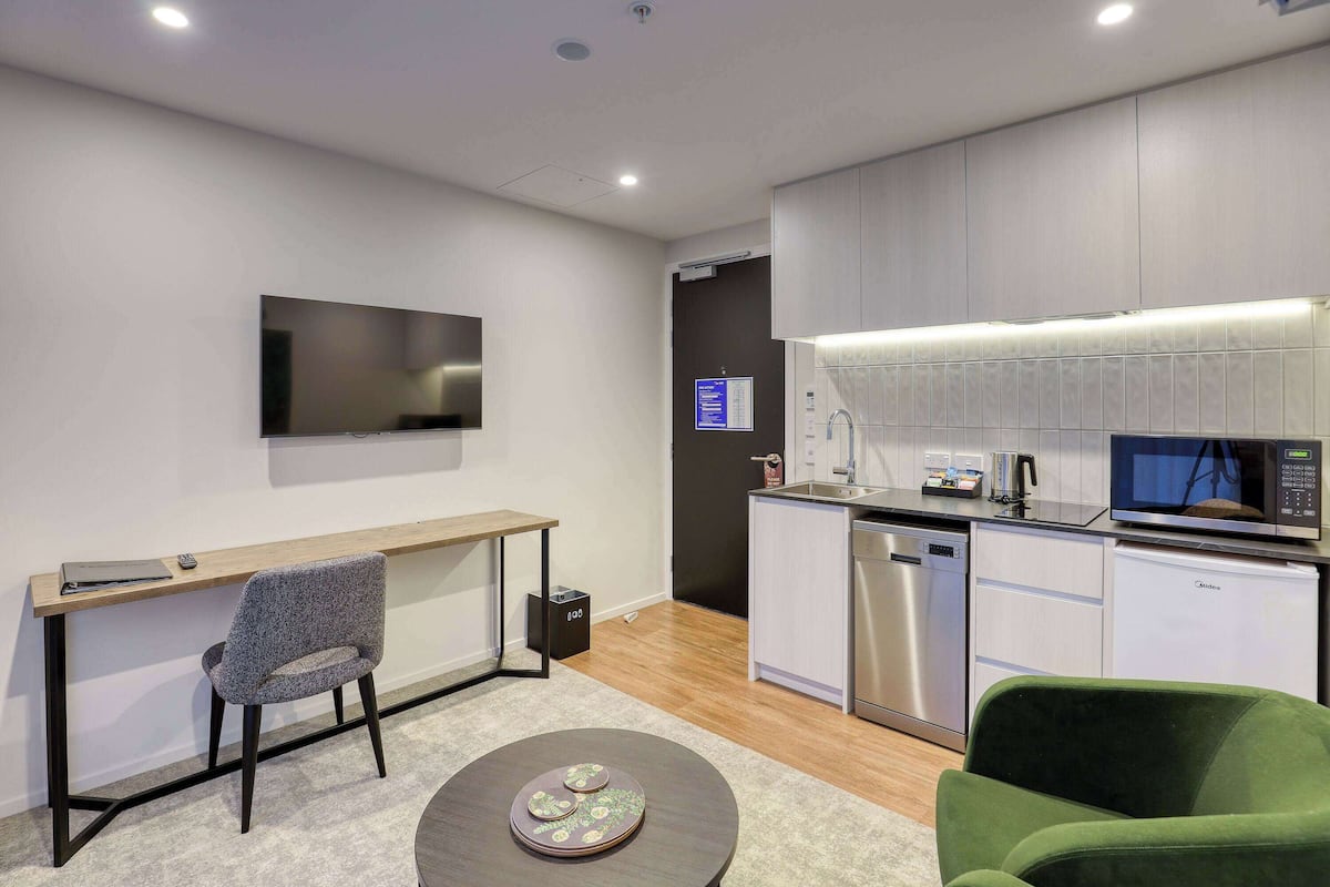 A modern kitchen with a flat-screen TV, a microwave, and a dishwasher. There is a desk with a chair and a small table with a decorative plate.