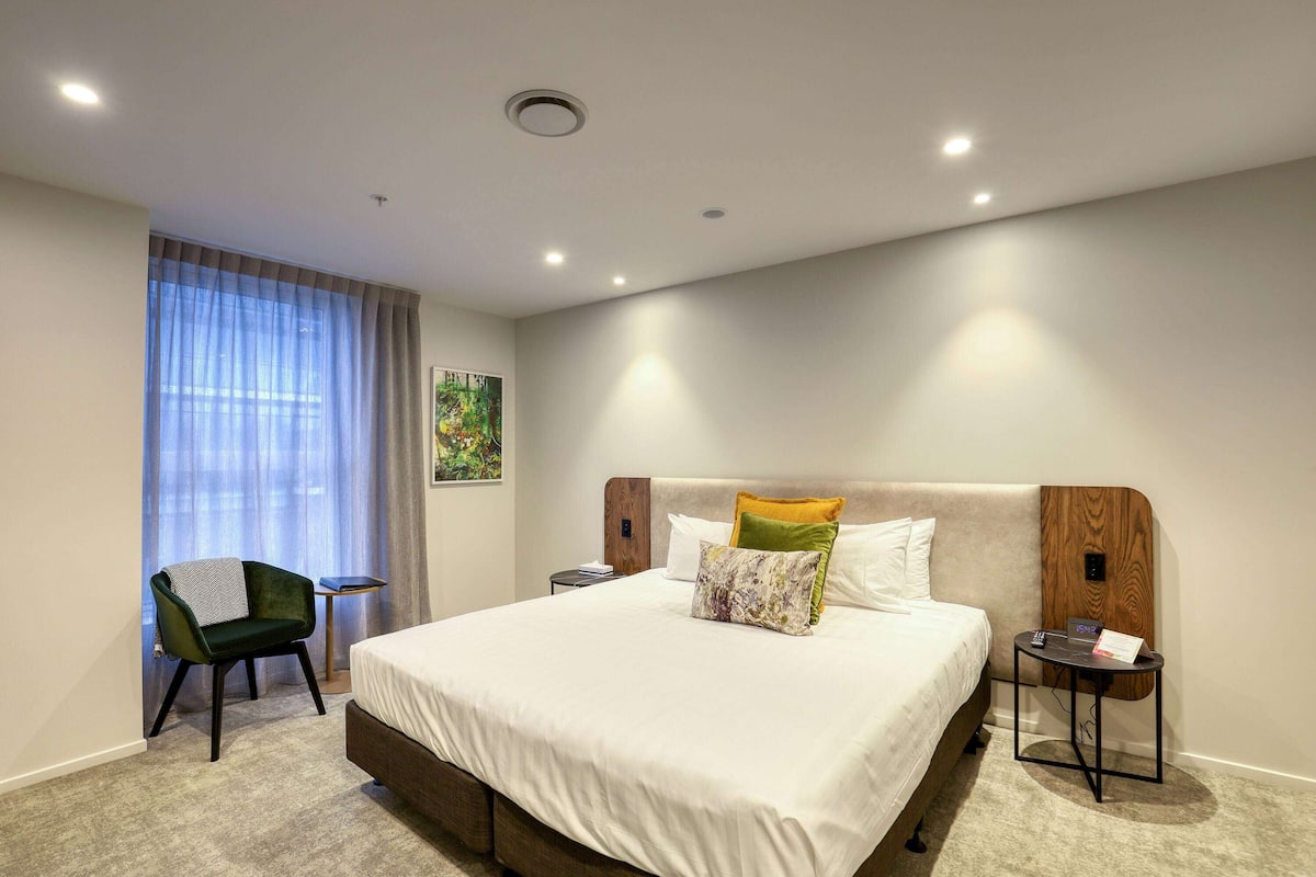 A modern hotel room with a large bed, a chair, a small table, and a nightstand.