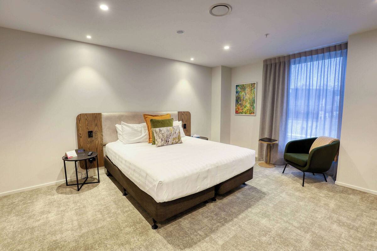 A modern hotel room with a large bed, a side table, a chair, and a window with curtains.