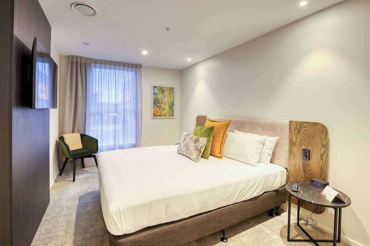 A hotel room with a large bed, a small table with a clock, a chair, and a TV mounted on the wall.