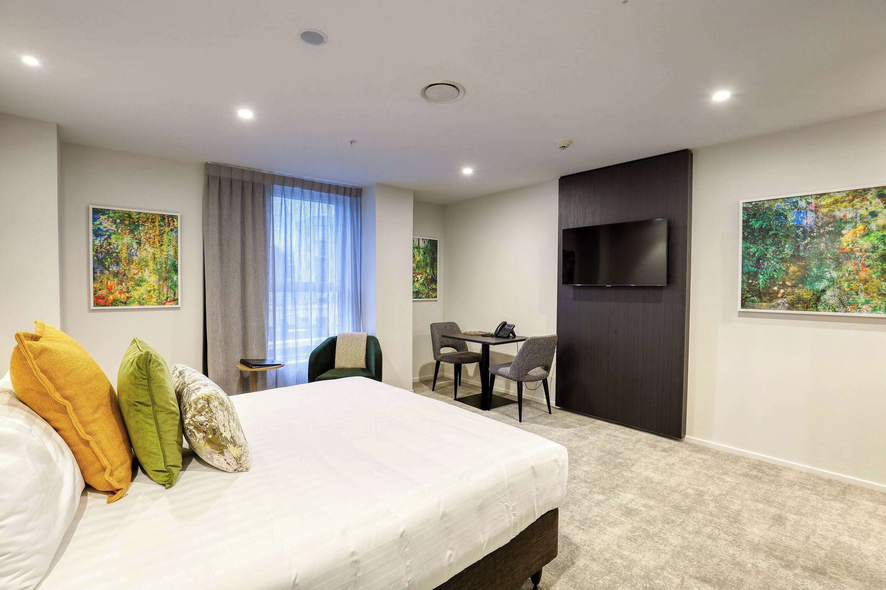 Habitación de hotel con una cama grande, un escritorio con dos sillas, un televisor de pantalla plana montado en la pared y dos cuadros en las paredes.
