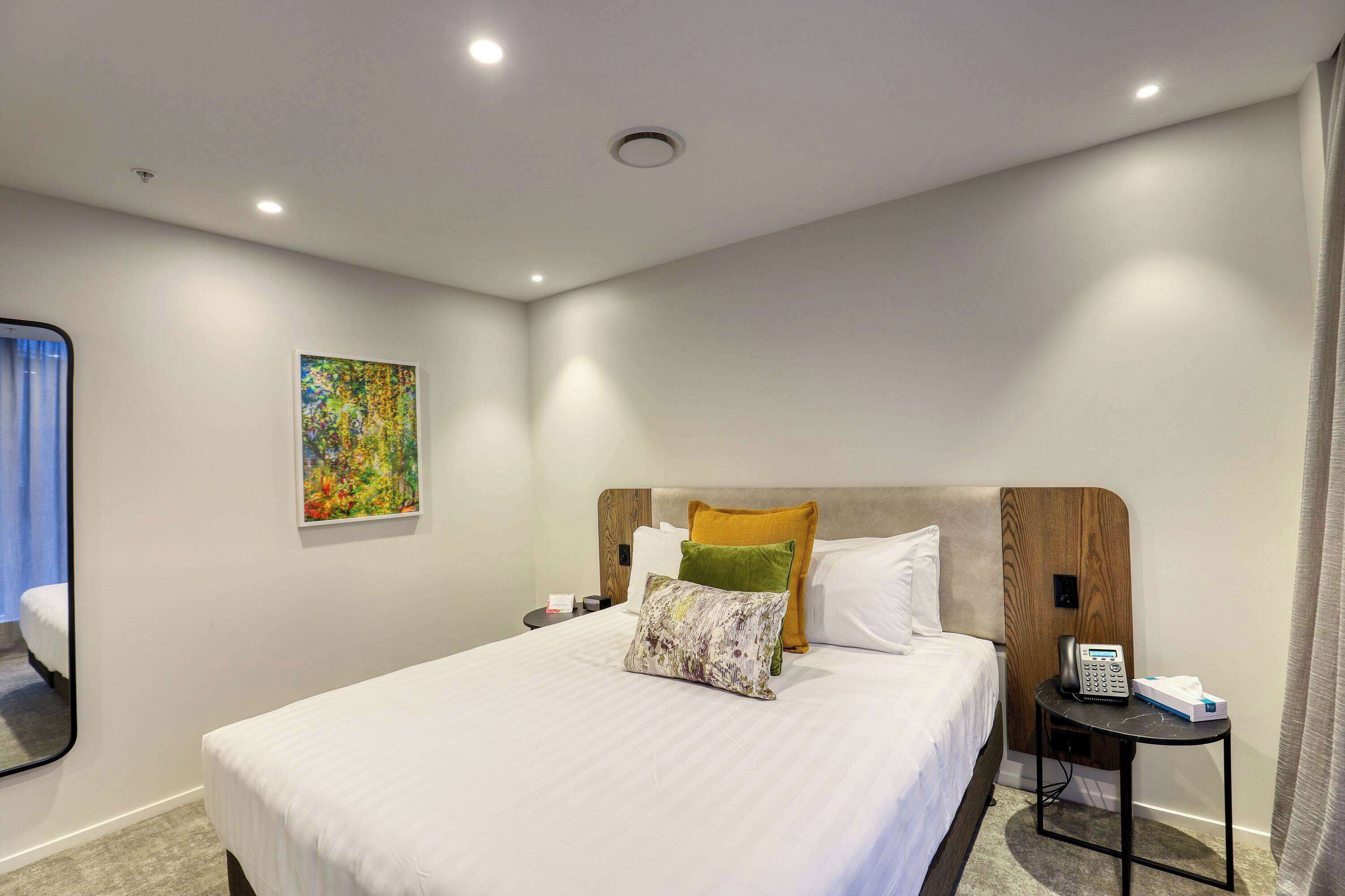 A hotel room with a large bed, a bedside table with a phone and tissues, a small side table, and a painting on the wall.