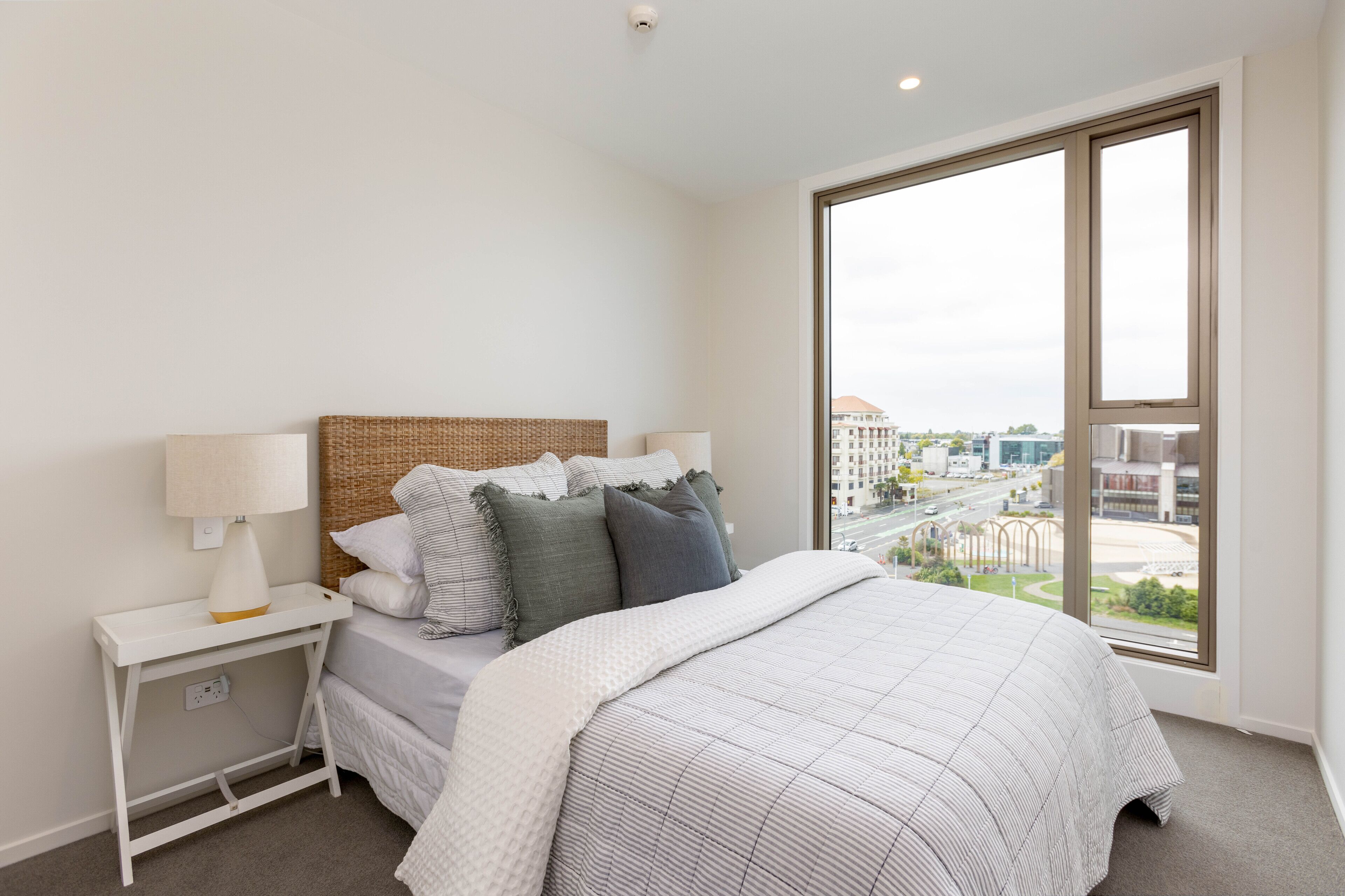 A modern bedroom with a large window, a bed with a patterned bedspread, a bedside table with a lamp, and a woven headboard.