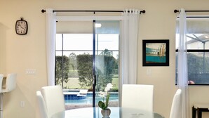 A dining area with a glass table and white chairs, a clock on the wall, and a view of a swimming pool and greenery outside. - Family vacation w/ Private Pool/Spa close to Disney,Parks & Golf (Champions gate)