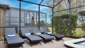A covered outdoor area with four lounge chairs, a hot tub, and a view of a building and trees. - Family vacation w/ Private Pool/Spa close to Disney,Parks & Golf (Champions gate)