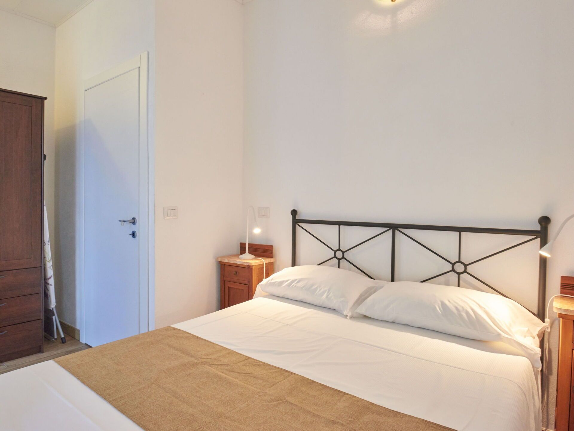 Een slaapkamer met een bed, houten nachtkastjes, een witte deur en een houten garderobe.