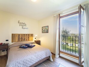 A bedroom with a bed, a bedside table, a nightstand, a wall-mounted headboard, a window with a view of greenery, and a decorative sign on the wall.