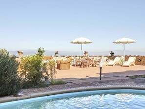 Un área de piscina en la azotea con sillones de descanso y sombrillas, rodeada por un muro de piedra y plantas en macetas.