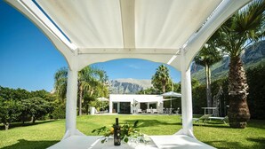 Een witte prieel met een gedekte tafel voor een huwelijksceremonie, met uitzicht op een modern huis met terras, omringd door weelderige begroeiing en palmbomen.