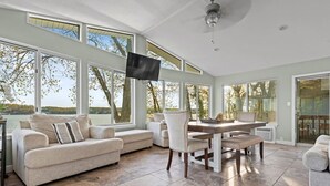 Un salon spacieux doté d’une grande fenêtre donnant sur un lac, un coin repas avec une table et des chaises, et un téléviseur à écran plat fixé au mur.