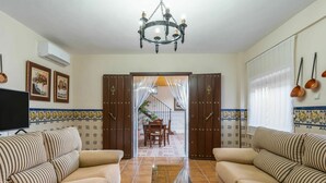 A living room with two sofas, a TV, and a chandelier. There is a dining area in the background with a table and chairs.