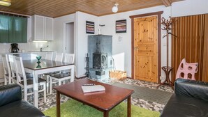 A cozy living room with a wooden ceiling, a dining area with white chairs, a black stove, and a wooden door.