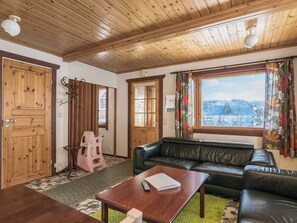 Een gezellige woonkamer met een houten plafond, een zwart leren bankstel, een houten salontafel en zicht op een besneeerd landschap door een raam.