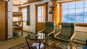 Una habitación acogedora con vigas de madera, una mesa de vidrio, dos sillas, una litera y una ventana con vistas nevadas.